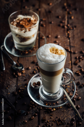 A glass of spicy latte with whipped cream and cinnamon, standing on a brown board. Coffee beans. Dark background