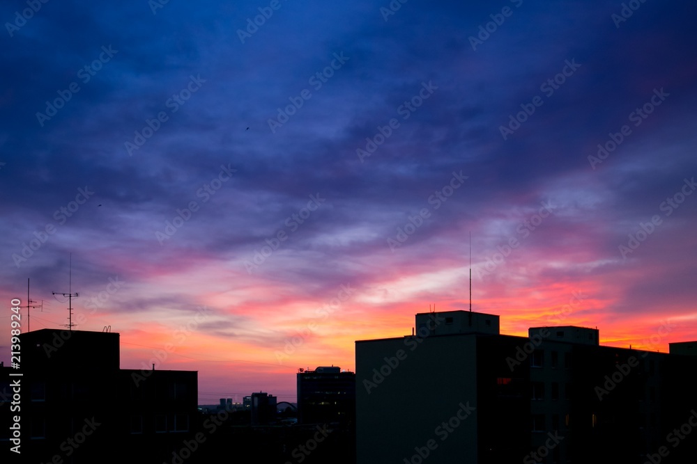 View of dark blue sky with clouds during sunset over black silhouettes of buildings, golden and pink colors behind houses, panorama, dusk landscape