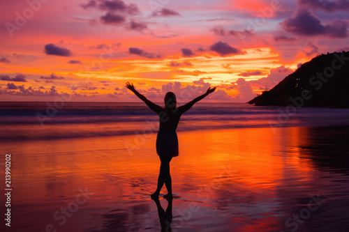 Silhouette of woman with hands up while standing on the sea beach at sunset.