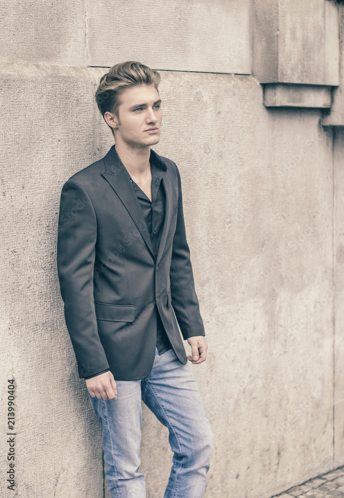 Handsome blond young man, blue eyes, leaning against white, rough stone wall, looking up to a side Handsome blond young man, blue eyes, leaning against white, rough stone wall, looking up to a side