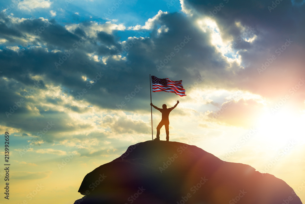 the man is standing on the top of the mountain, holding his hands up with the flag of the America against the sunset. Business concept idea, success and achievements, career ladder, victory.