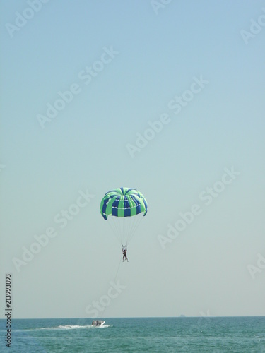 The man was attached to a long line to a moving boat. On a blue-green parachute, a man soars in the air. Theme active recreation in the resort, parasailing, having fun in summer
