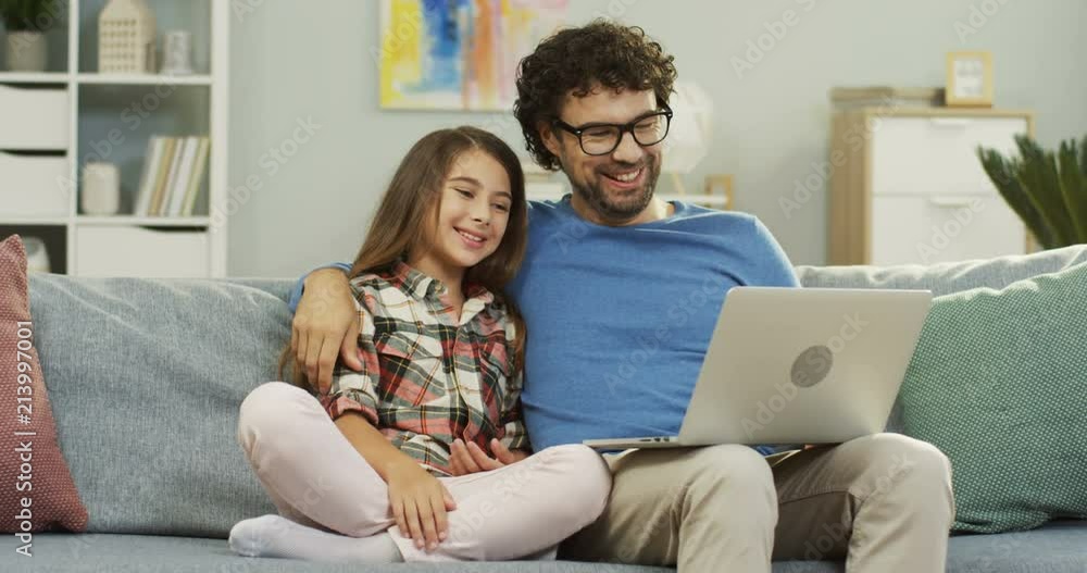 Portrait shot of the handsome father in glasses hugging his pretty little daughter while they having videochat on the laptop computer at home. Indoors. Portrait shot of the handsome father in glasses hugging his pretty little daughter while they having videochat on the laptop computer at home. Indoors.