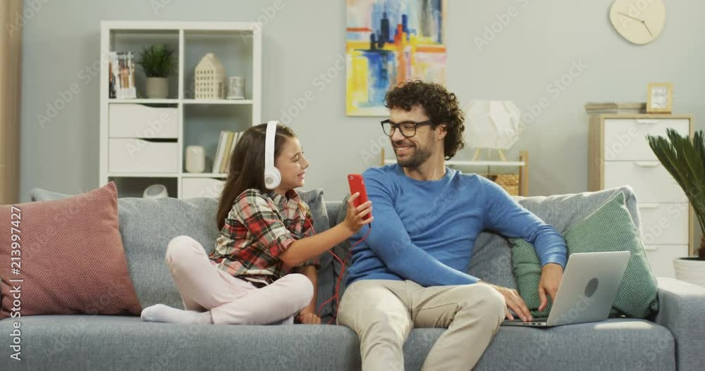 Caucasian attractive man sitting on the couch and using laptop computer while his small daughter in headphones using smartphone and showing something to the dad. Inside. Caucasian attractive man sitting on the couch and using laptop computer while his small daughter in headphones using smartphone and showing something to the dad. Inside.