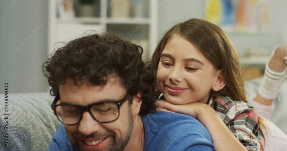 Close up of the smiled good looking father in glasses lying on the couch with his small cute daughter on his back and they watching something on the tablet. Indoor. Close up of the smiled good looking father in glasses lying on the couch with his small cute daughter on his back and they watching something on the tablet. Indoor.