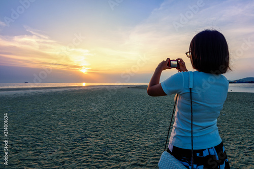 Asian woman tourist taking photo on the beach and beautiful natural landscape of colorful sky and sea during a sunset at Nathon Sunset Viewpoint in Samui island, Surat Thani, Thailand