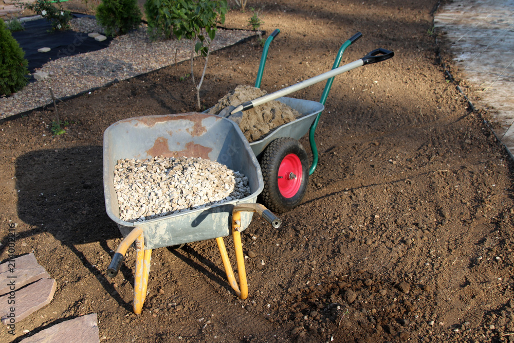 Wheelbarrows loaded with crushed stone, sand and a square point shovel on the cultivated soil in