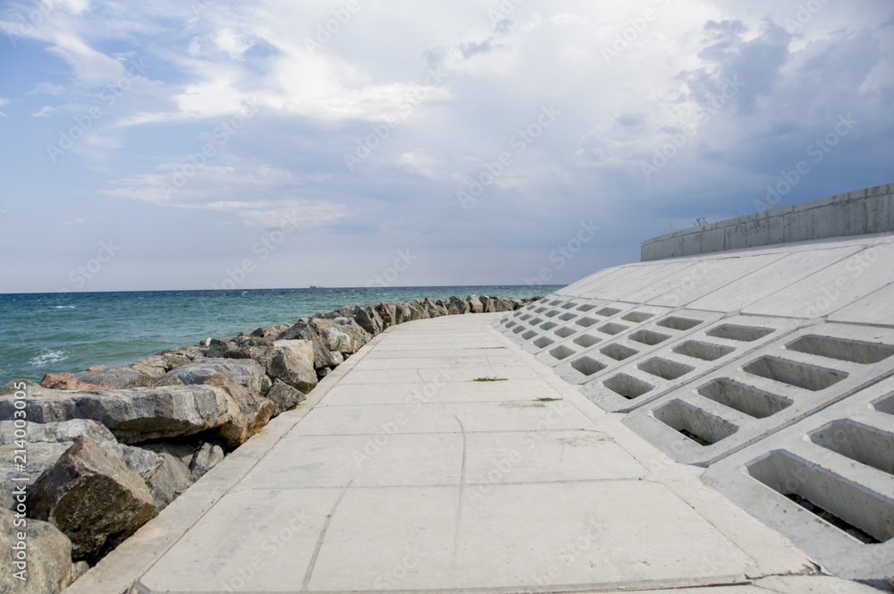 Background of the sea wave in summer. Water landscape with clouds on the horizon. Sea nature of tranquility. An alarming sky. Stones by the water, a concrete path along the sea.