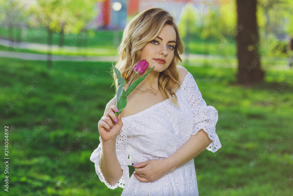 Young beautiful blonde girl in white dress with a flower tulip on a background of green grass Young beautiful blonde girl in white dress with a flower tulip on a background of green grass