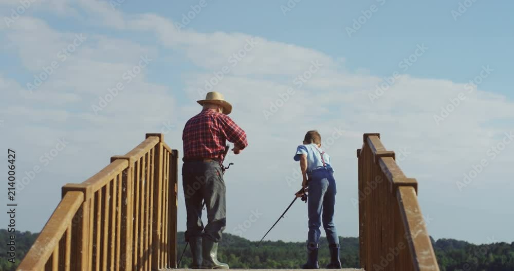 Back view on the little boy pulling out a fish while he fishing together with his grandpa on the bridge. Outdoor.