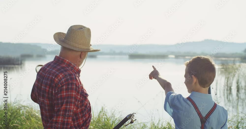 Rear of the grandfather and grandson holding their fishings rods and talking on the river bank. Outdoor. Rear of the grandfather and grandson holding their fishings rods and talking on the river bank. Outdoor.