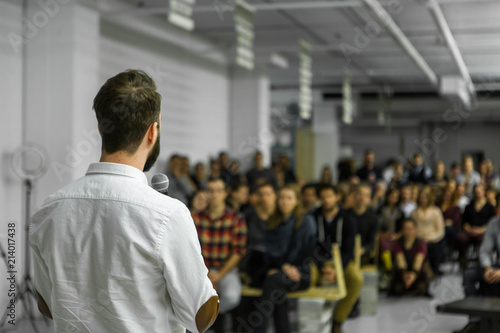Man with beard gives a public speech in front of 200 people, in an industrial environment