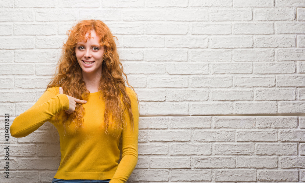 Young redhead woman standing over brick wall with surprise face pointing finger to himself Young redhead woman standing over brick wall with surprise face pointing finger to himself