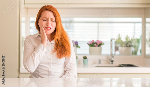 Redhead woman at kitchen touching mouth with hand with painful expression because of toothache or dental illness on teeth. Dentist concept.