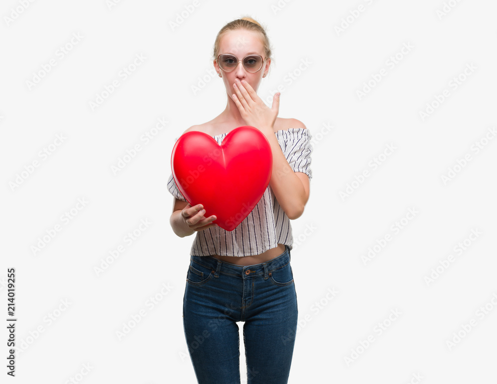 Blonde teenager woman holding red heart cover mouth with hand shocked with shame for mistake, expression of fear, scared in silence, secret concept Blonde teenager woman holding red heart cover mouth with hand shocked with shame for mistake, expression of fear, scared in silence, secret concept