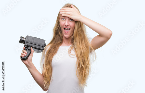 Blonde woman teenager filming holding super 8 video camera stressed with hand on head, shocked with shame and surprise face, angry and frustrated. Fear and upset for mistake.
