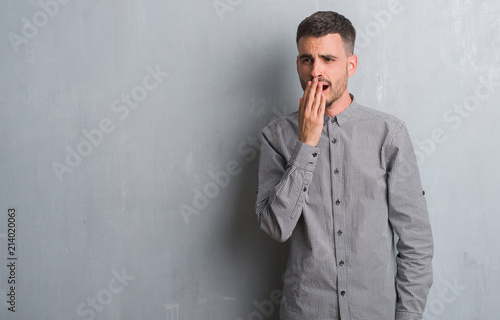 Young adult man standing over grey grunge wall bored yawning tired covering mouth with hand. Restless and sleepiness.