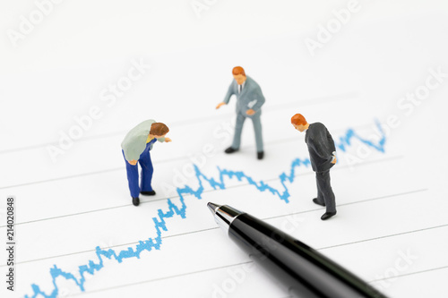 Financial analysis, investment consultant or advisor concept, miniature professional businessmen standing and looking, review on volatility blue stock market line graph data report with pen
