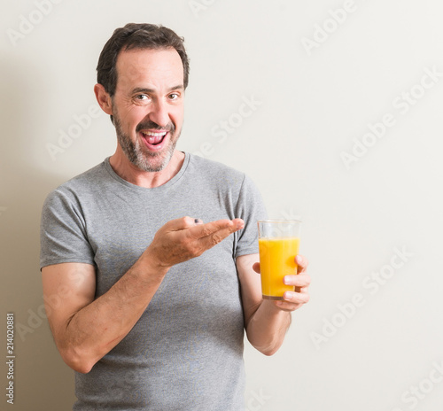 Senior man drinking orange juice in a glass very happy pointing with hand and finger