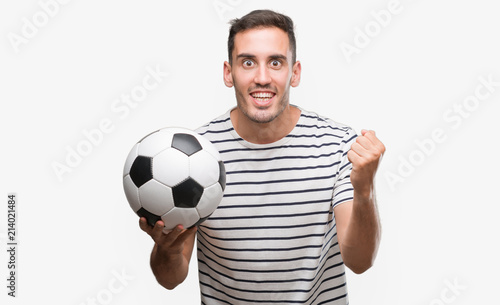 Handsome young man holding soccer football screaming proud and celebrating victory and success very excited, cheering emotion