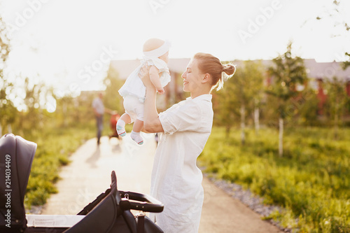 Young mother walks in the park in the evening with baby. Mom took out her daughter from the carriage and laughs at her with a pleasant smile. Sunset, nature, happiness, family