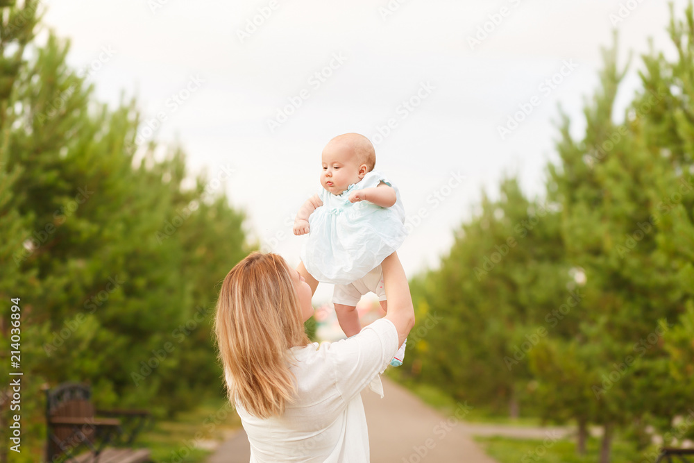 Fototapeta premium Happy young mom and adorable toddler playing together in park in summertime, smiling mother holding her little daughter lifted up, looking at baby with love and adoration surrounded by trees. Family