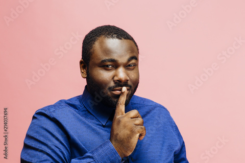 Don't tell anyone! Young man in blue shirt holding finger over his mouth, isolated on pink background