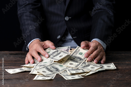 Pile of paper currency ..Man hands in pinstripe suit sweeping pile of US dollar banknotes on old wooden table and black background,business concept.
