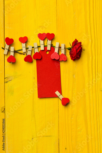 Congratulation and wishes concept. Flower with blank tag or card on yellow wooden background. Tag for congratulation pinned to flower, copy space. Rose flower with tiny pins with hearts on stem