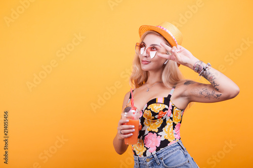 Charming young woman making peace signs while having a fresh orange juice with sunglasses over yellow bacground in studio
