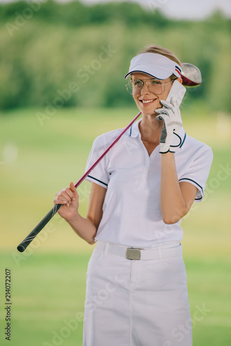 portrait of smiling female golf player in polo and cap with golf club in hand talking on smartphone at golf course