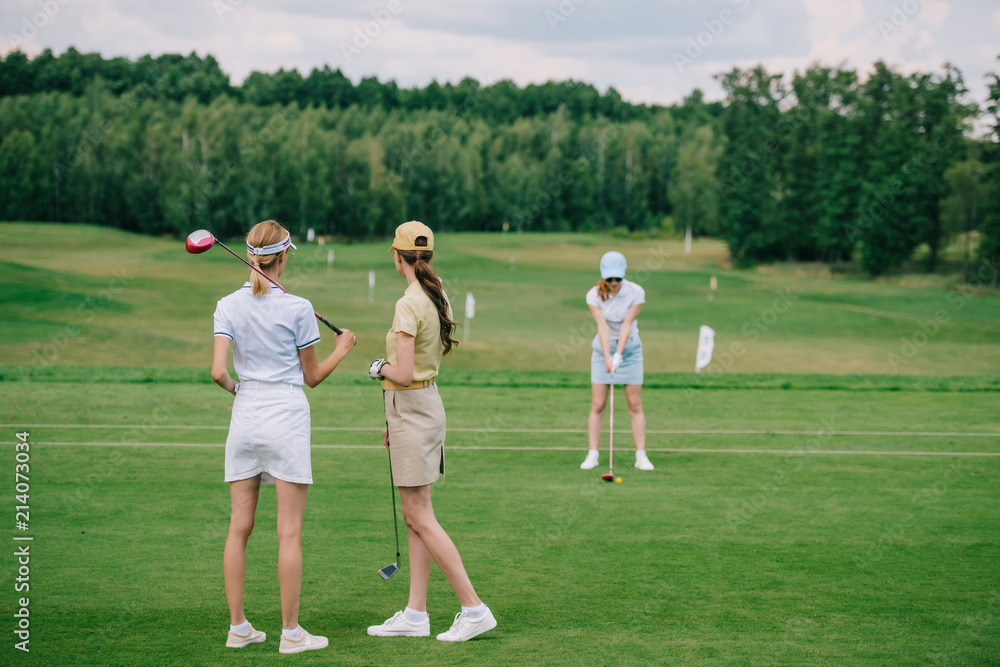 selective focus of women in caps with golf equipment looking at friend playing golf at golf course selective focus of women in caps with golf equipment looking at friend playing golf at golf course