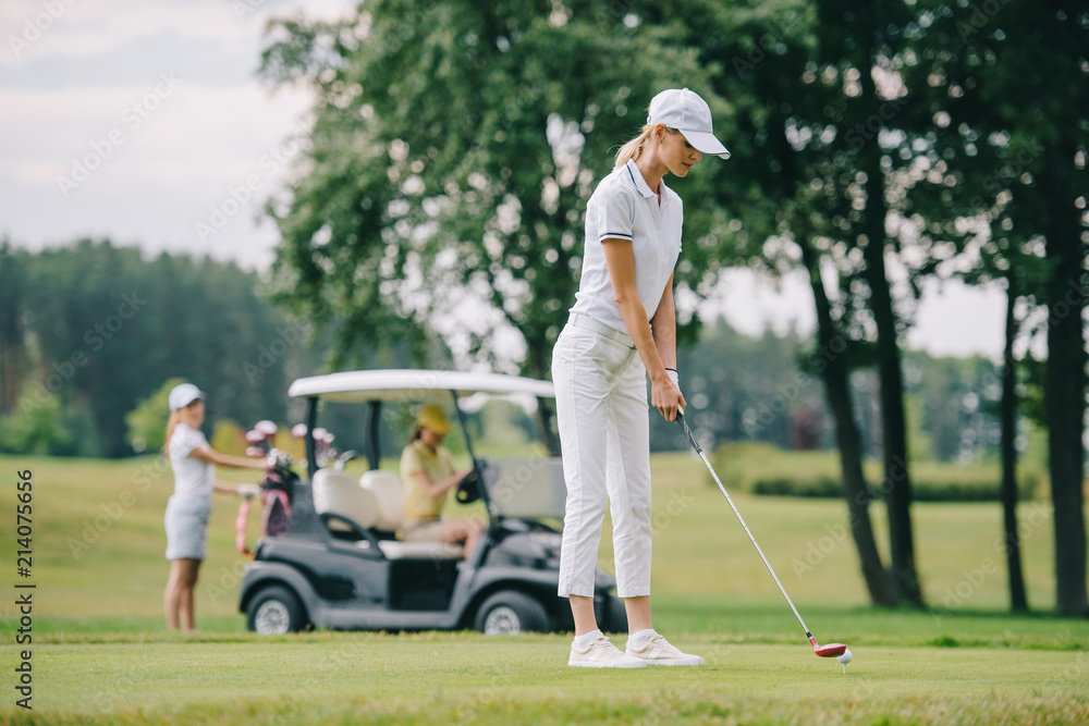 Obraz premium selective focus of woman with golf club playing golf and friends resting at golf cart on green lawn