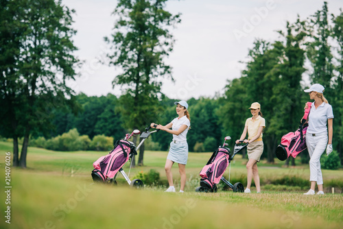 Smiling women in caps with golf equipment walking on golf course