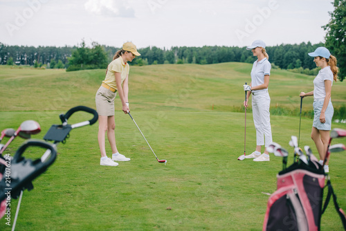 side view of woman in cap playing golf while friends standing near by at golf course
