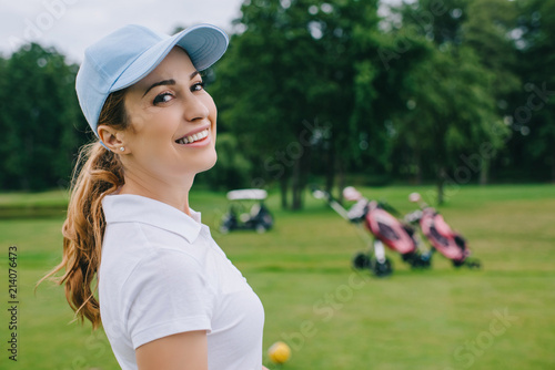 side view of smiling woman in polo and cap looking at camera at golf course
