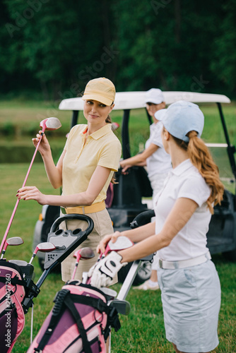 selective focus of female golf players with golf equipment and friend in golf cart behind on green lawn