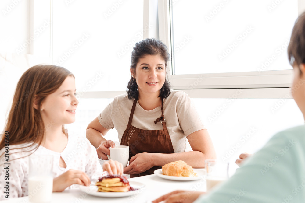 Photo of cheerful family woman and little daughter sitting at table in bright kitchen, and eating pancakes Photo of cheerful family woman and little daughter sitting at table in bright kitchen, and eating pancakes