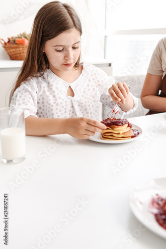 Portrait of beautiful ittle daughter 8-10 sitting at table in bright kitchen, and eating pancakes with jam
