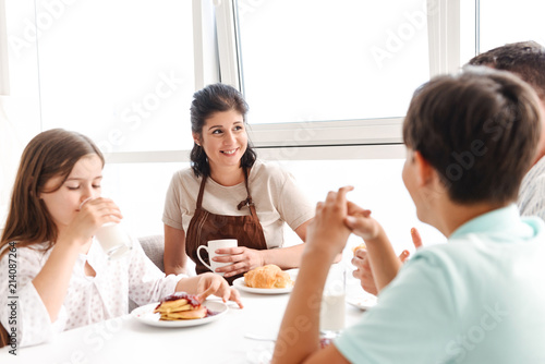 Photo of smiling parents with children 8-10 having breakfast together in bright kitchen at home, while eating croissants and pancakes