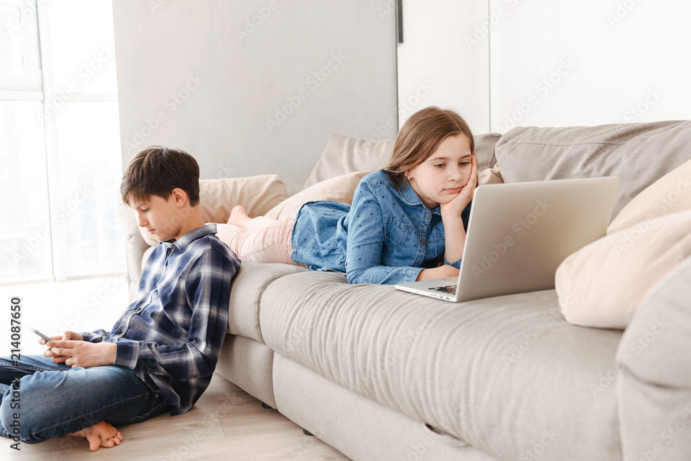 Image of two modern european children boy and girl 8-10 resting on sofa at home, while using mobile phone and laptop Image of two modern european children boy and girl 8-10 resting on sofa at home, while using mobile phone and laptop