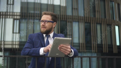 Businessman in glasses and blue suit reading busines news on brand-new tablet standing near modern office building. Outdoora. Portrait. photo