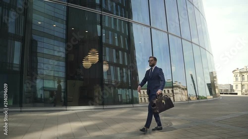Young stylish businessman is walking along street on modern building background, rich business owner with leather bag in hand, is going confidently, looking at watch in summer day. Concept