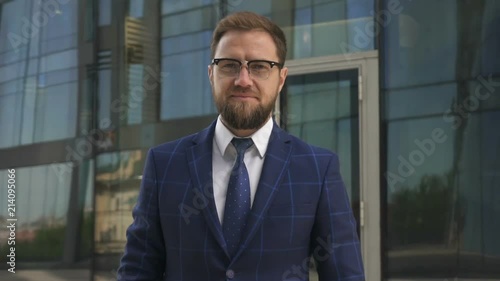 Confinet businessman with beard, in glasses and blue suit taking awat his brand new smartphone, looking to the camera and smiling. Outdoors. Portrait. photo