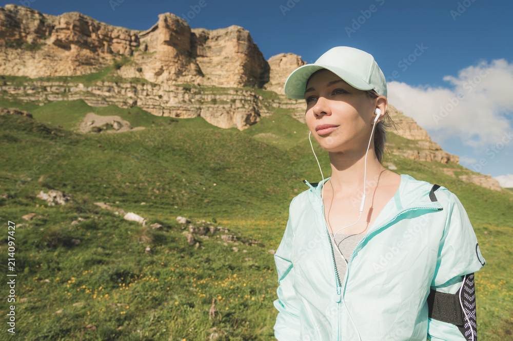 Naklejka premium Fitness runner woman listening to music on the nature. Portrait of beautiful girl wearing earphones earbuds and running cap. against the background of rocks field and blue sky