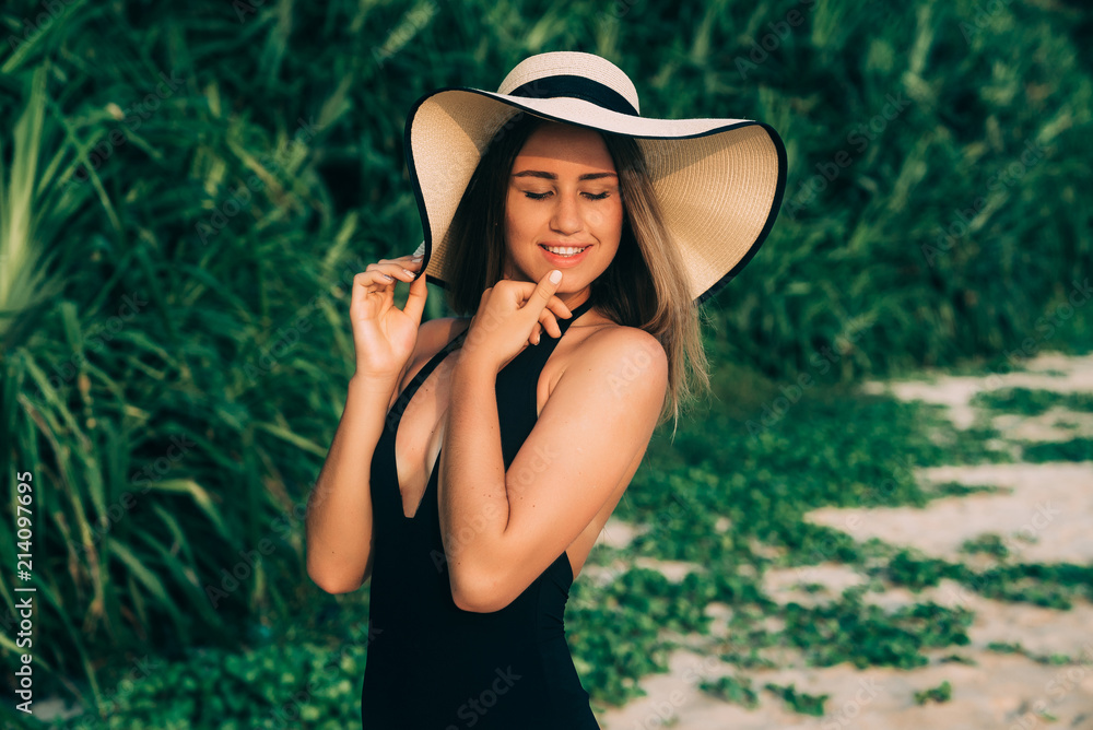 Portrait of a beautiful young girl, has a smooth tanned young skin. Enjoys the holiday, wears a swimsuit and a stylish beach hat, closes her eyes with pleasure, playfully touches her lips