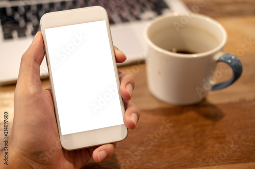 Mockup image of a man's hand holding white mobile phone with blank desktop screen with coffee cup and laptop on wooden table