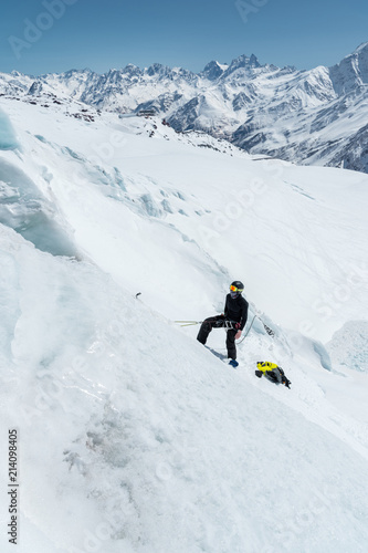 A professional mountaineer in a helmet and ski mask on the insurance does notch the ice ax in the glacier. The work of a professional climber in winter on a glacier against the blue sky
