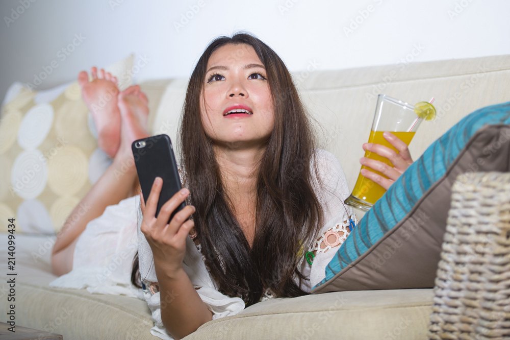 young beautiful and happy Asian Chinese woman on her 20s or 30s lying at living room sofa couch drinking healthy orange juice using internet mobile young beautiful and happy Asian Chinese woman on her 20s or 30s lying at living room sofa couch drinking healthy orange juice using internet mobile