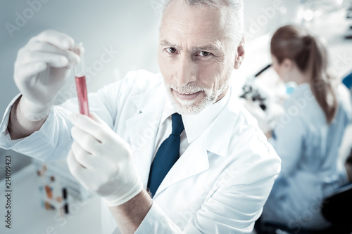 Chemical experiment. Smart nice experienced chemist holding a test tube and looking at it while working in the lab